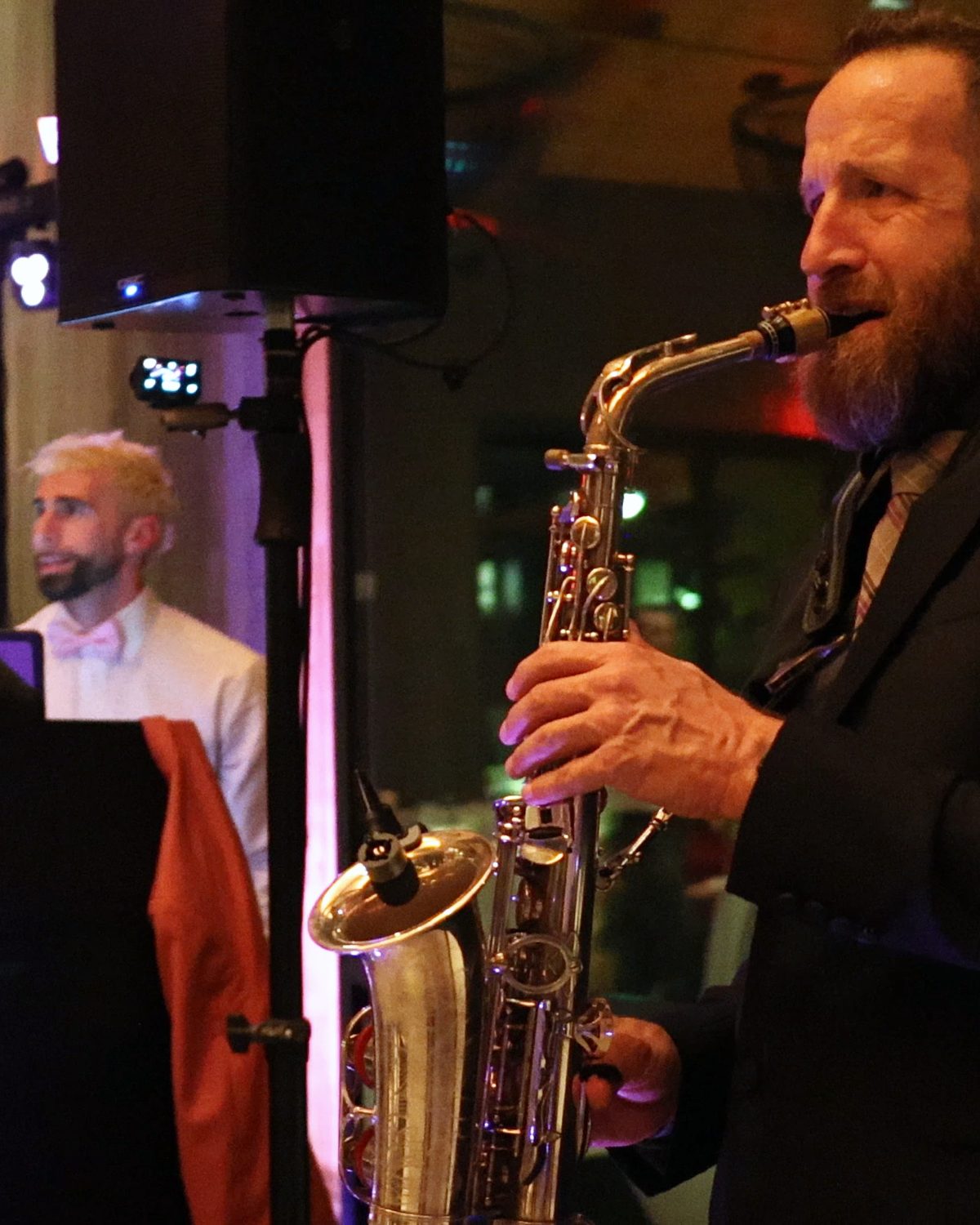 A saxophonist plays in the foreground while DJ Shazam reads the room from the booth behind him