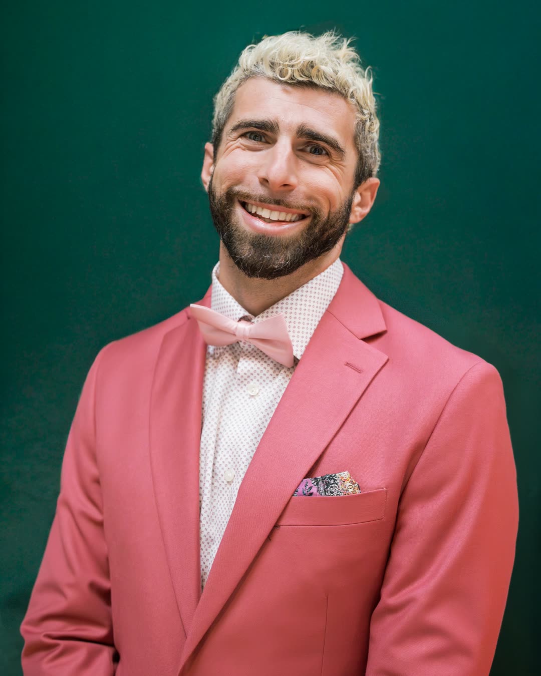 DJ Shazam, smiling, in a coral jacket against an evergreen backdrop