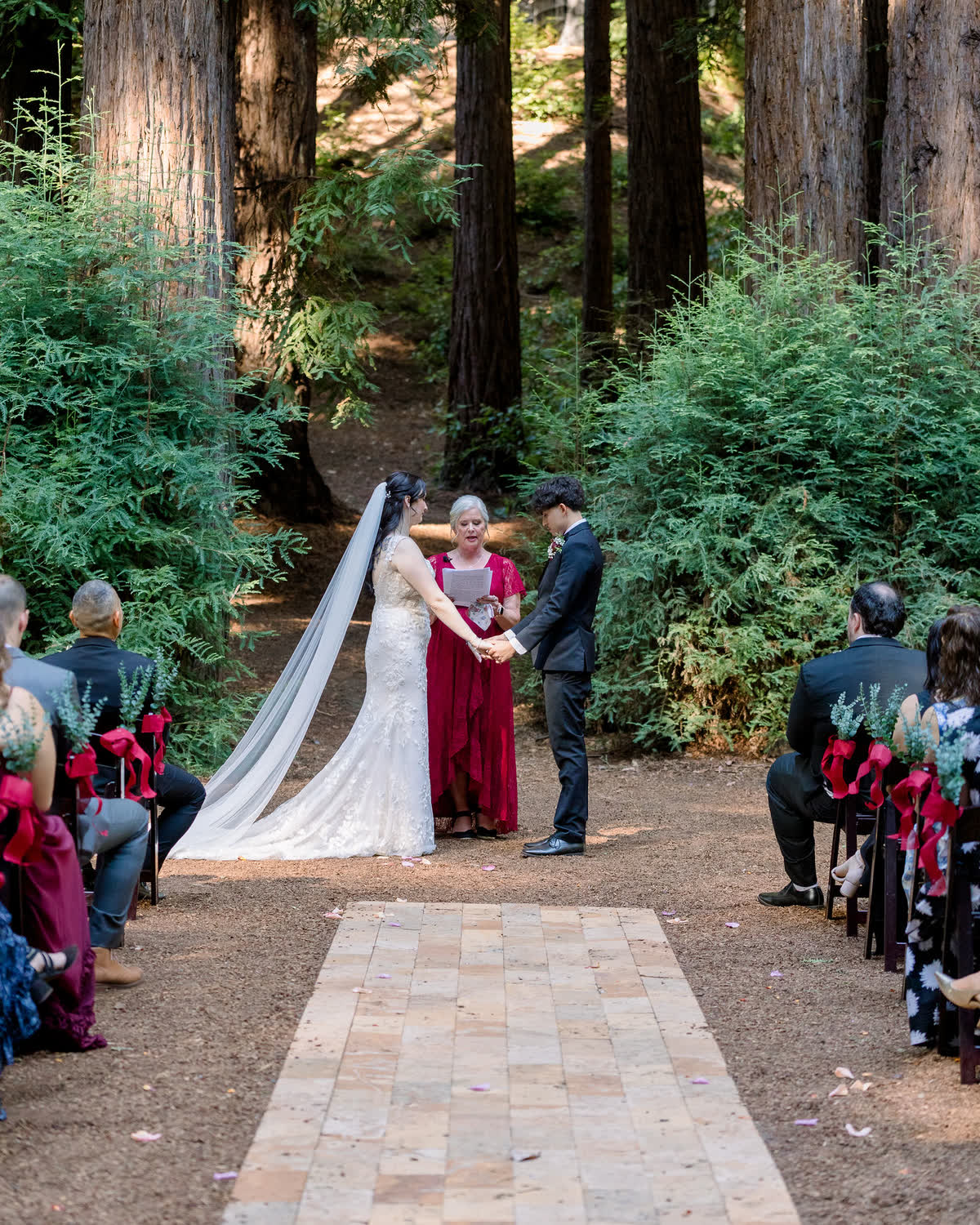 A couple holding hands at the altar in the Waterfall Lodge redwood grove, the officiant between them and guests seated along the brick aisle